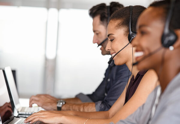 Smiling multiethnic people with headsets using computer and smiling while working in office. Young man and woman operators talking on headset with clients. Group of telemarketing customer service team working while sitting in a row.