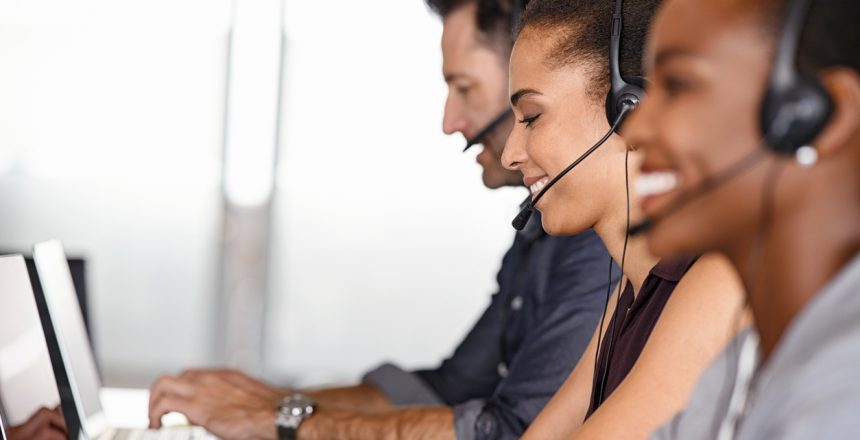 Smiling multiethnic people with headsets using computer and smiling while working in office. Young man and woman operators talking on headset with clients. Group of telemarketing customer service team working while sitting in a row.